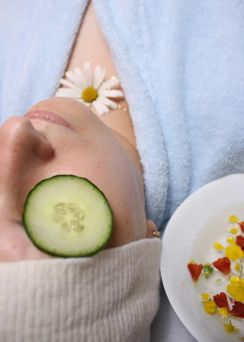 A person relaxing with a cucumber slice on their eye, a daisy on their chest, and a plate of colorful flower petals beside them A person relaxing with a cucumber slice on their eye, a daisy on their chest, and a plate of colorful flower petals beside them