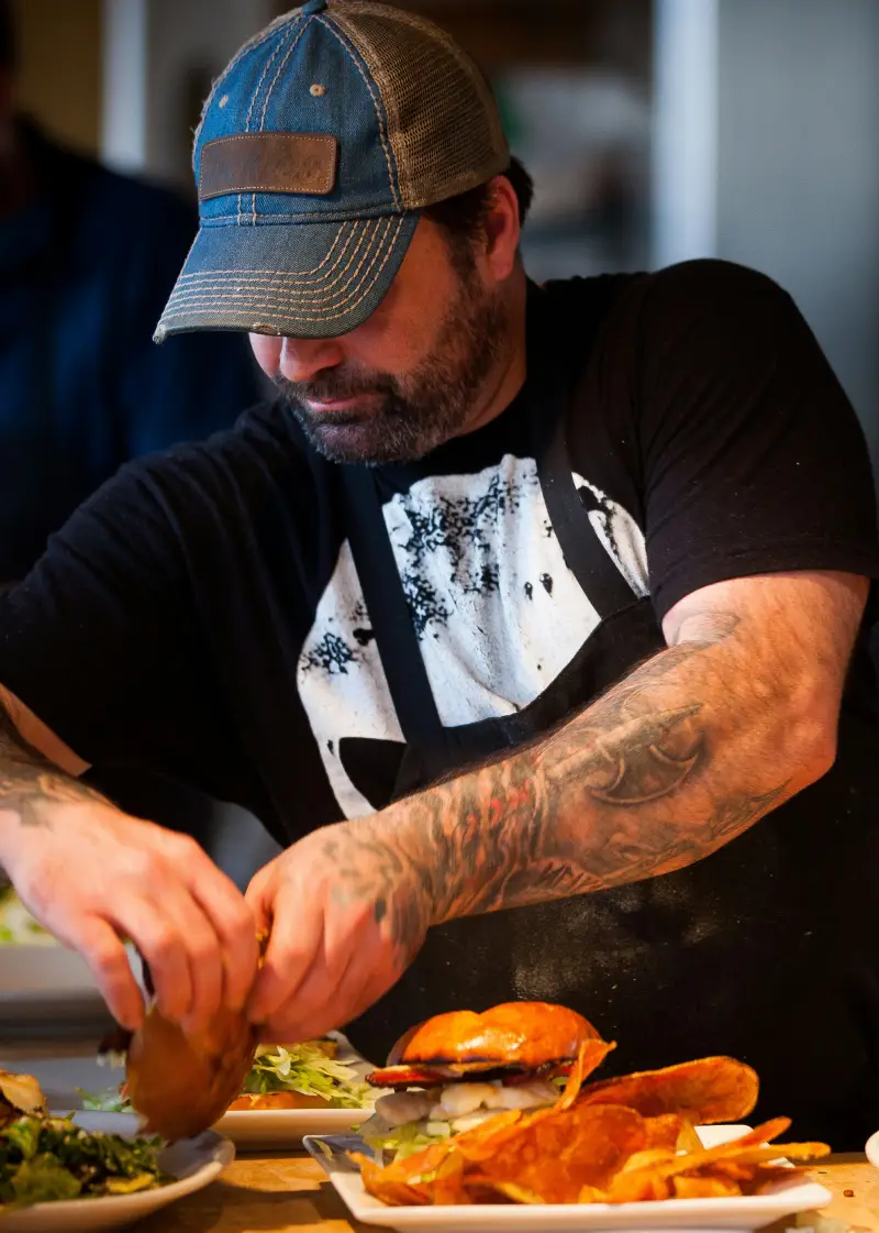 A tattooed chef prepares a plate with a burger, chips, and salad in a lively kitchen setting, showcasing culinary skills and creativity A tattooed chef prepares a plate with a burger, chips, and salad in a lively kitchen setting, showcasing culinary skills and creativity